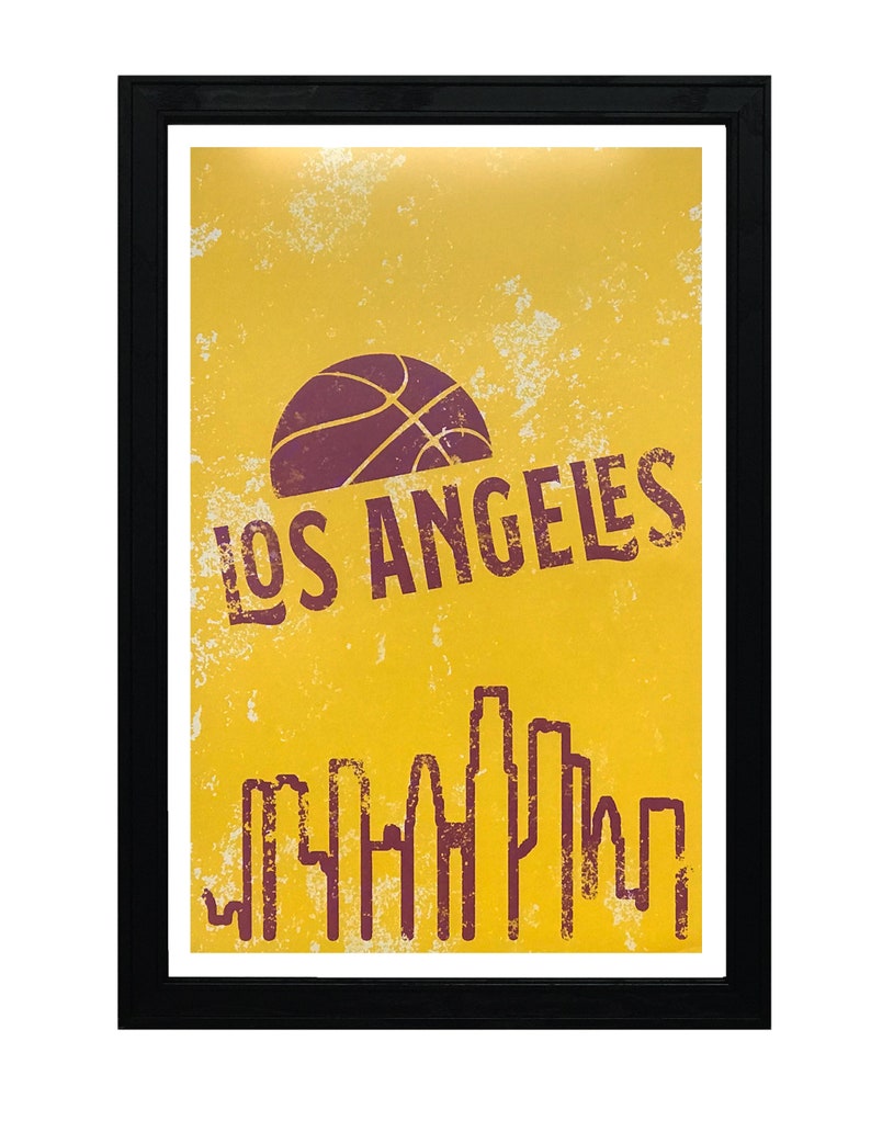 May include: A framed poster with a yellow background and a distressed texture. The poster features a basketball graphic, the text "LOS ANGELES", and a skyline silhouette in a dark red color. The frame is black.