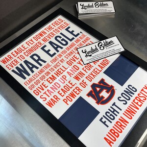 K&ouml;nnte beinhalten: Ein gerahmter Druck mit wei&szlig;em Hintergrund und schwarzem Rahmen. Der Druck zeigt ein blaues und oranges Logo der Auburn University mit dem Text "War Eagle, Fly Down the Field, Ever to Conquer, Never to Yield." und "Fight Song, Auburn University" in rot und blau.