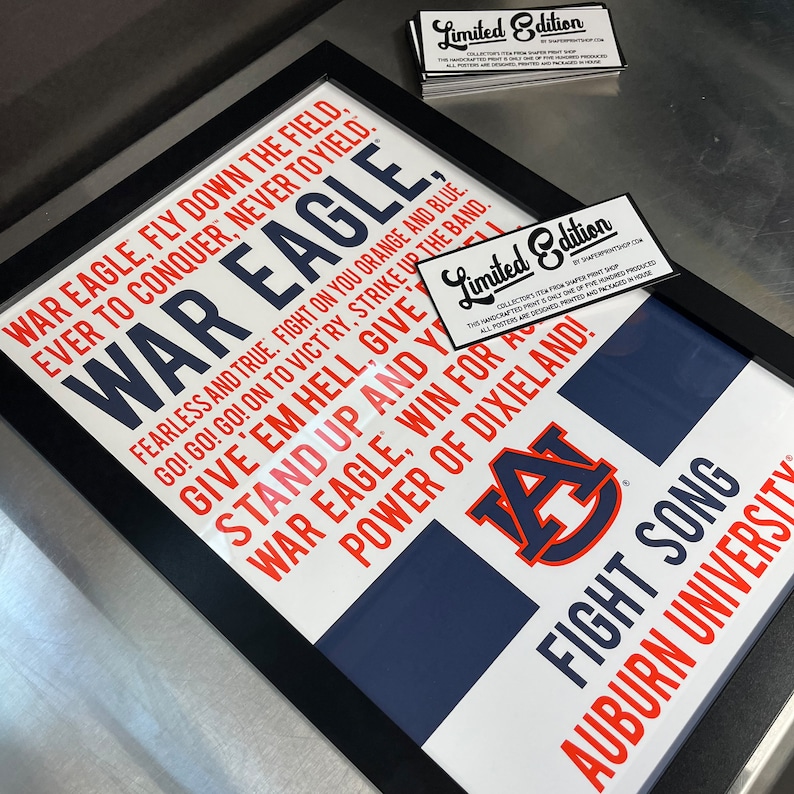 K&ouml;nnte beinhalten: Ein gerahmter Druck mit wei&szlig;em Hintergrund und schwarzem Rahmen. Der Druck zeigt ein blaues und oranges Logo der Auburn University mit dem Text "War Eagle, Fly Down the Field, Ever to Conquer, Never to Yield." und "Fight Song, Auburn University" in rot und blau.