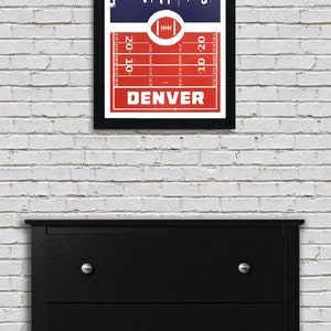 May include: A framed print of a red and blue football field with a white silhouette of the Denver skyline. The text "DENVER" is printed in white at the bottom of the field.
