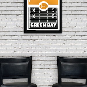 May include: A black framed poster with a yellow and black football field design. The poster features a white silhouette of the Green Bay skyline and the text "GREEN BAY". The poster is hanging on a white brick wall between two black bar stools.