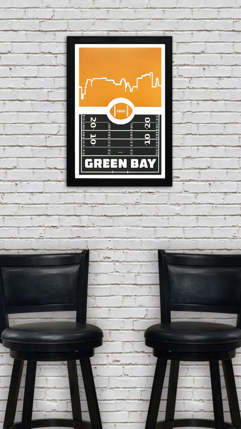 May include: A black framed poster with a yellow and black football field design. The poster features a white silhouette of the Green Bay skyline and the text "GREEN BAY". The poster is hanging on a white brick wall between two black bar stools.