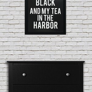 May include: Black and white framed print with the text "I like my coffee black and my tea in the harbor". The print is hanging on a white brick wall above a black dresser with four drawers.