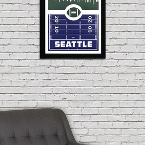 May include: A framed print of a football pitch with a skyline of Seattle, Washington. The pitch is blue with white lines and the skyline is green with white buildings. The text "SEATTLE" is printed in white at the bottom of the print.