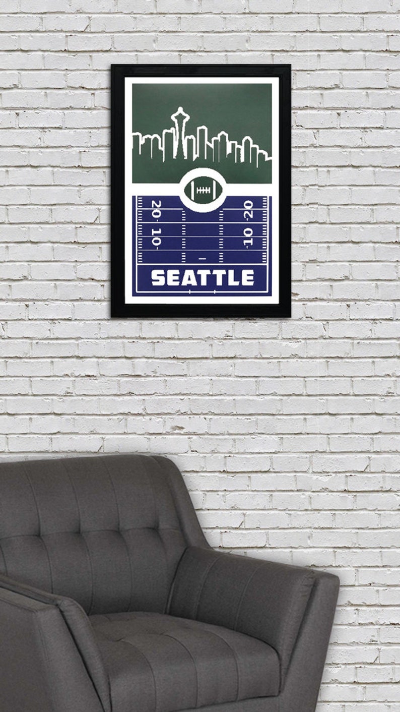 May include: A framed print of a football pitch with a skyline of Seattle, Washington. The pitch is blue with white lines and the skyline is green with white buildings. The text "SEATTLE" is printed in white at the bottom of the print.