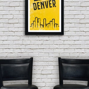 Puede incluir: Un p&oacute;ster enmarcado en negro con fondo amarillo y un gr&aacute;fico de baloncesto. El texto "DENVER" est&aacute; en letras negras en el p&oacute;ster. El p&oacute;ster est&aacute; colgado en una pared de ladrillo blanco. Dos taburetes de bar negros est&aacute;n frente a la pared.