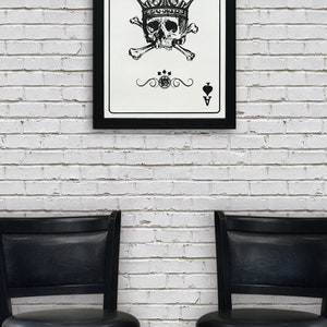 May include: A black framed print of a playing card with a skull wearing a crown and crossed bones. The card is the Ace of Spades. The print is hanging on a white brick wall between two black bar stools.