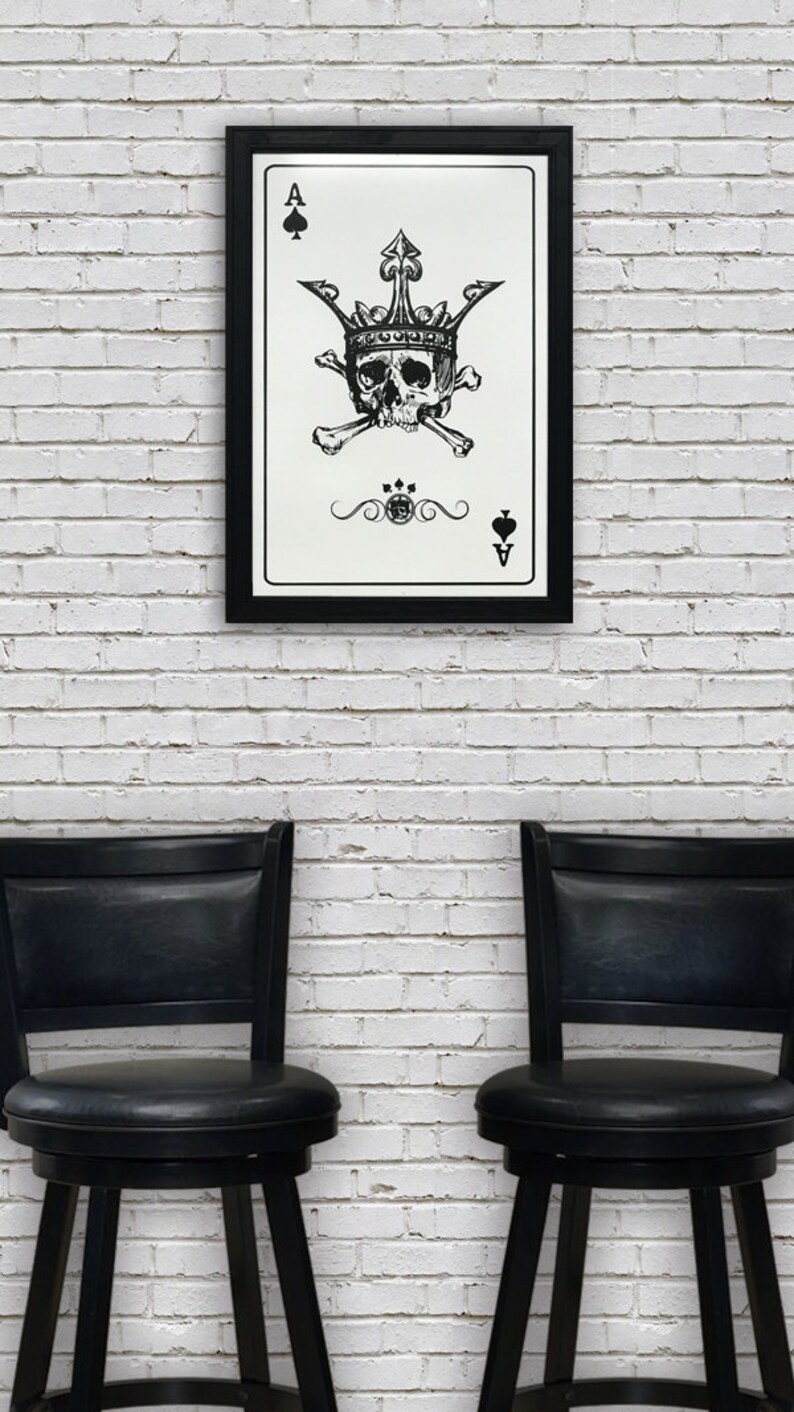 May include: A black framed print of a playing card with a skull wearing a crown and crossed bones. The card is the Ace of Spades. The print is hanging on a white brick wall between two black bar stools.