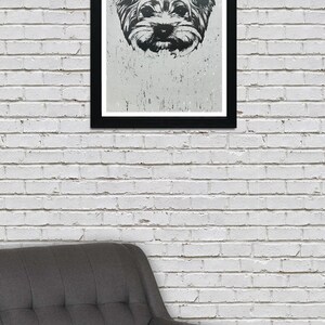 May include: A black and white framed print of a Yorkshire Terrier dog's face. The dog is looking directly at the viewer with a serious expression. The print is hanging on a white brick wall above a gray armchair.