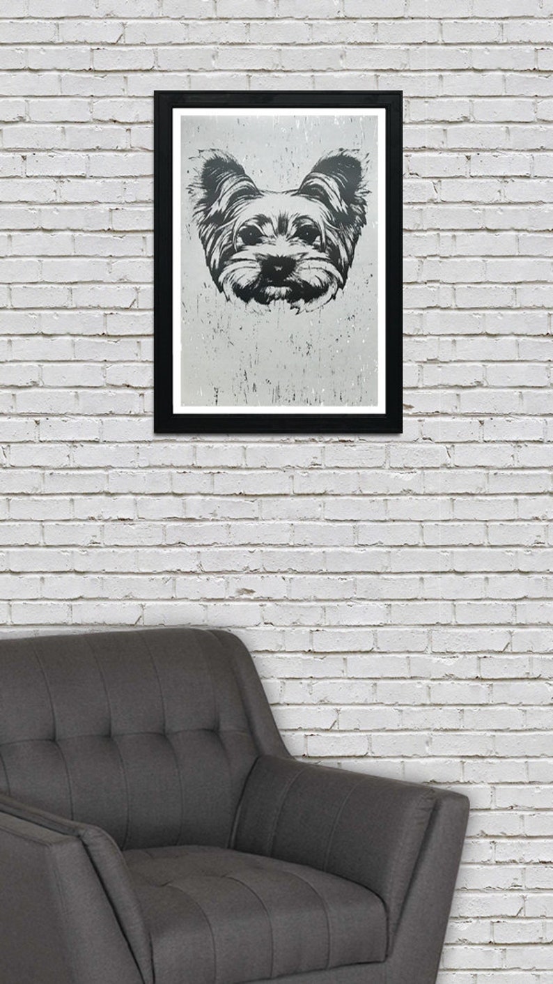 May include: A black and white framed print of a Yorkshire Terrier dog's face. The dog is looking directly at the viewer with a serious expression. The print is hanging on a white brick wall above a gray armchair.