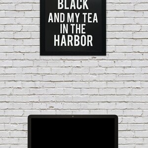 May include: A black framed sign with white text that reads "I like my coffee black and my tea in the harbor" hanging on a white brick wall. A laptop computer with a black screen is on a dark wood surface in front of the wall.
