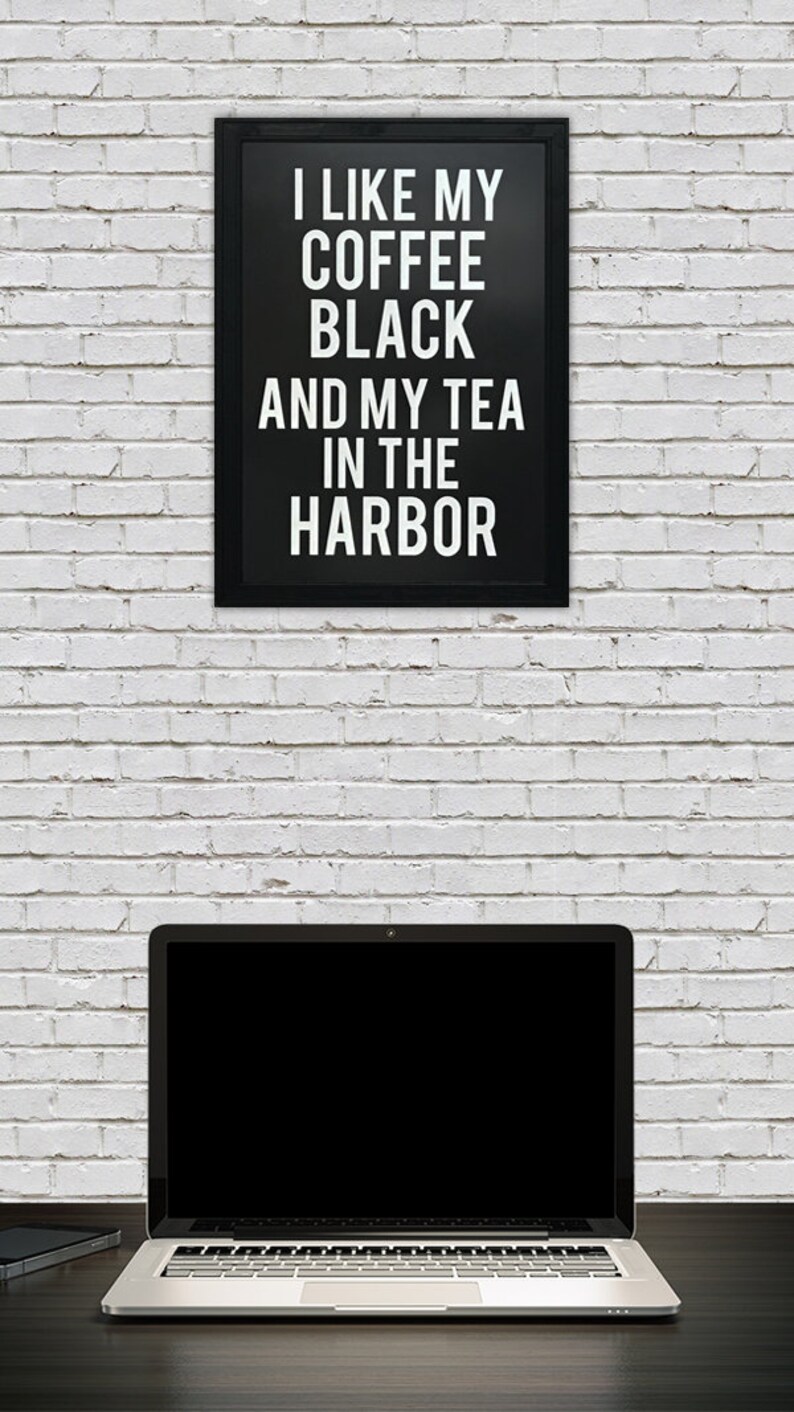 May include: A black framed sign with white text that reads "I like my coffee black and my tea in the harbor" hanging on a white brick wall. A laptop computer with a black screen is on a dark wood surface in front of the wall.