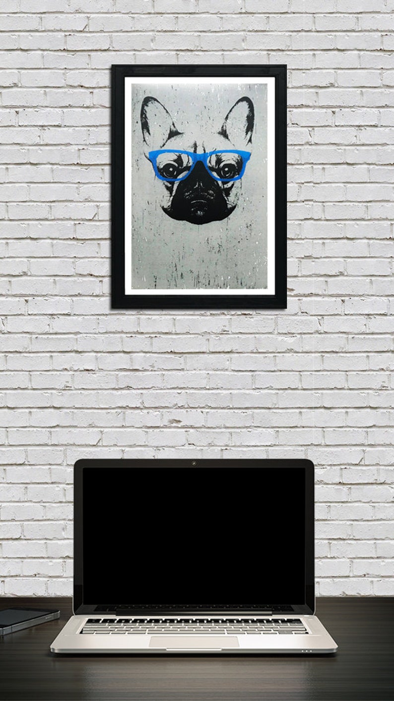 May include: A black and white illustration of a French bulldog wearing blue glasses. The illustration is framed in a black frame and hanging on a white brick wall. A laptop computer is sitting on a dark surface in front of the framed illustration.