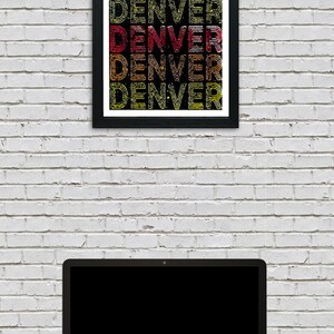 May include: A framed print with the word "DENVER" repeated in a stacked pattern. The letters are outlined in black and filled with a gradient of red, orange, and yellow. The print is hanging on a white brick wall, with a laptop computer in the foreground.