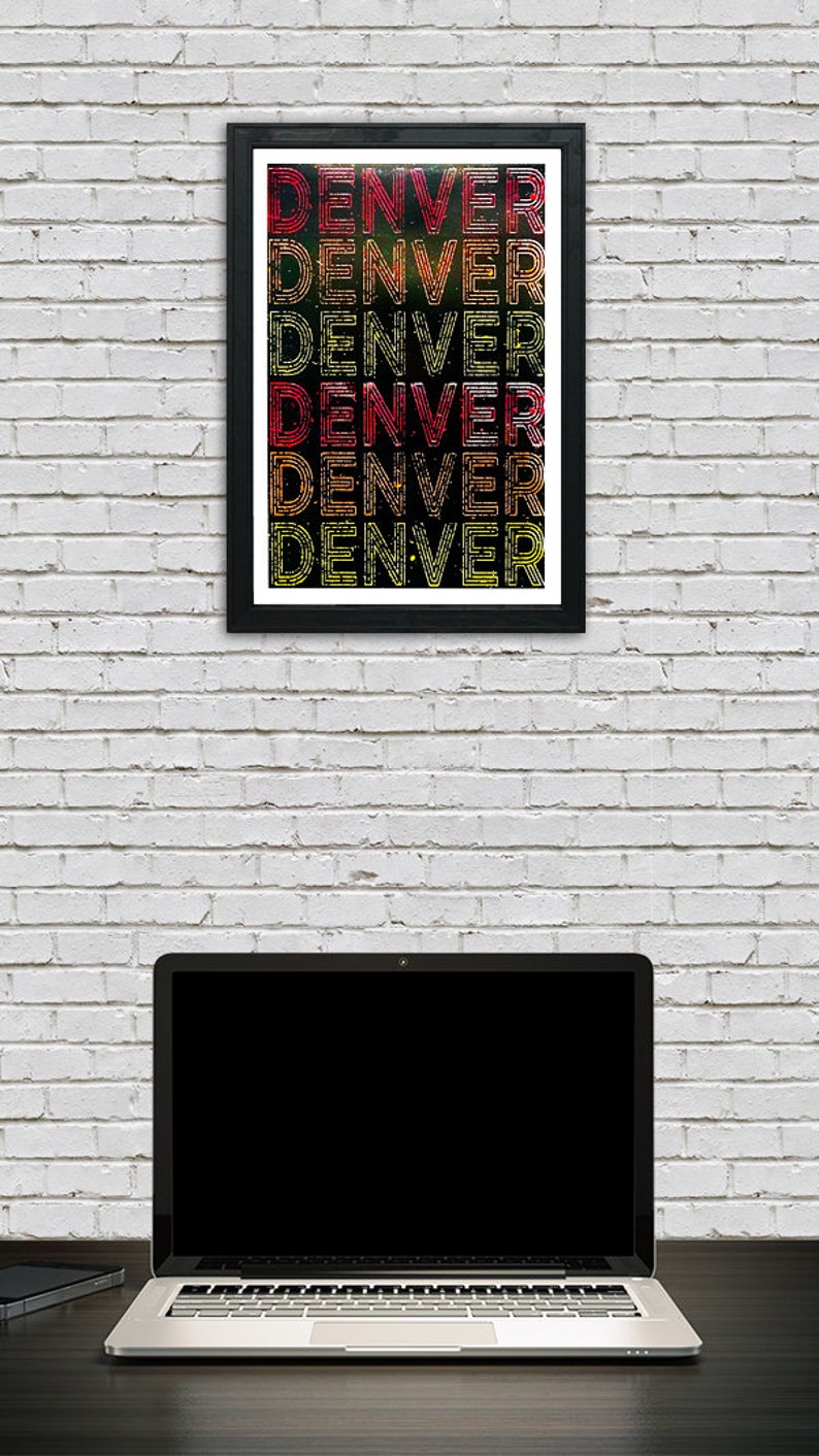 May include: A framed print with the word "DENVER" repeated in a stacked pattern. The letters are outlined in black and filled with a gradient of red, orange, and yellow. The print is hanging on a white brick wall, with a laptop computer in the foreground.
