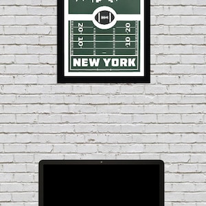 Puede incluir: Una impresi&oacute;n enmarcada verde y blanca con una ciudad estilizada y un dise&ntilde;o de campo de f&uacute;tbol. El texto "NEW YORK" est&aacute; impreso debajo del campo. La impresi&oacute;n est&aacute; colgada en una pared de ladrillo blanco.