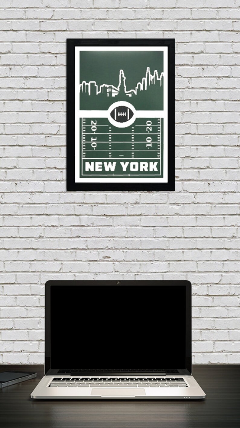Puede incluir: Una impresi&oacute;n enmarcada verde y blanca con una ciudad estilizada y un dise&ntilde;o de campo de f&uacute;tbol. El texto "NEW YORK" est&aacute; impreso debajo del campo. La impresi&oacute;n est&aacute; colgada en una pared de ladrillo blanco.