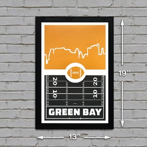 May include: A black framed print with a yellow cityscape silhouette and a black and white football field graphic. The text "GREEN BAY" is printed in white on the field. The print measures 13 inches wide by 19 inches tall.