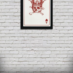 May include: Framed art print featuring a red Ace of Hearts card design. The card shows a skull wearing a crown, with crossbones beneath it. The background is white, and the frame is black. A pile of poker chips is in the foreground.