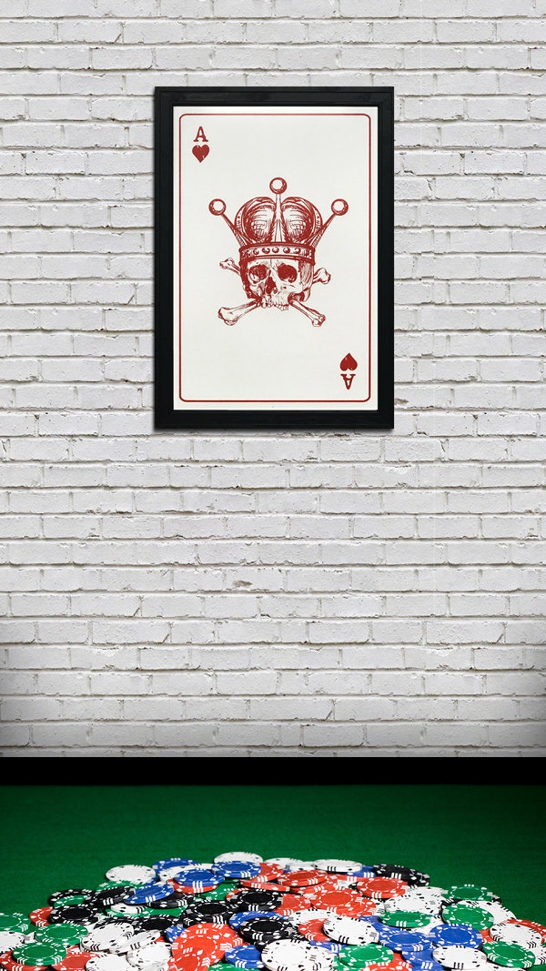 May include: Framed art print featuring a red Ace of Hearts card design. The card shows a skull wearing a crown, with crossbones beneath it. The background is white, and the frame is black. A pile of poker chips is in the foreground.