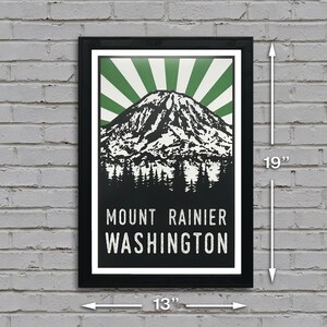 May include: A black framed poster with a green and white sunburst design behind a black and white illustration of Mount Rainier. The text "Mount Rainier Washington" is printed in white at the bottom of the poster. The poster measures 13 inches wide by 19 inches tall.
