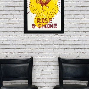 May include: A framed print with a yellow background and a rooster illustration. The rooster is red and yellow and is standing on a yellow sunburst. The text "RISE & SHINE" is printed in red below the rooster.