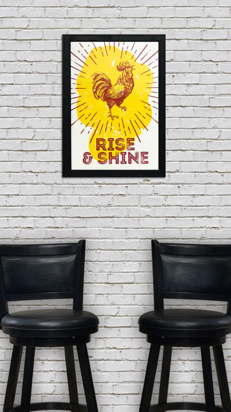 May include: A framed print with a yellow background and a rooster illustration. The rooster is red and yellow and is standing on a yellow sunburst. The text "RISE & SHINE" is printed in red below the rooster.