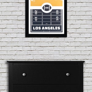 May include: A framed print with a black background and a white outline of the Los Angeles skyline. The skyline is on a yellow background. Below the skyline is a football field with yard lines and a football in the center. The text "LOS ANGELES" is below the football field.