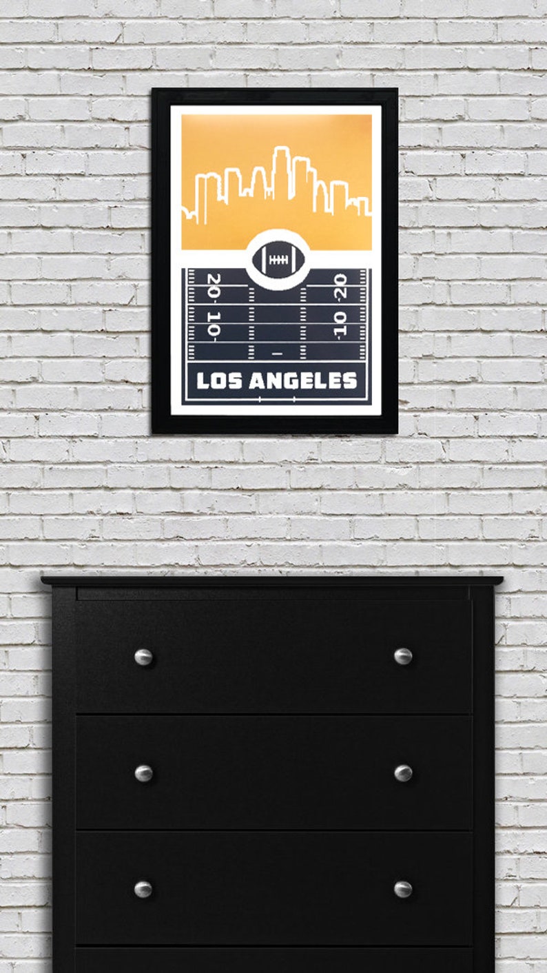 May include: A framed print with a black background and a white outline of the Los Angeles skyline. The skyline is on a yellow background. Below the skyline is a football field with yard lines and a football in the center. The text "LOS ANGELES" is below the football field.