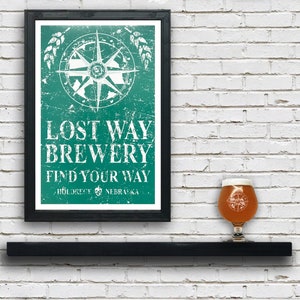 May include: A framed poster with a distressed green background featuring a compass and the text "Lost Way Brewery Find Your Way  Hildrece NEBRASKA". A glass of beer with a compass design is sitting on a black shelf in front of the poster.