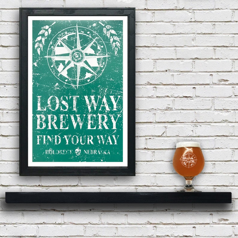 May include: A framed poster with a distressed green background featuring a compass and the text "Lost Way Brewery Find Your Way  Hildrece NEBRASKA". A glass of beer with a compass design is sitting on a black shelf in front of the poster.