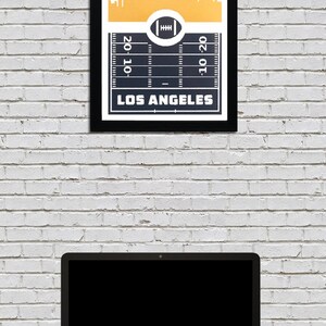 May include: A framed print with a gold skyline of Los Angeles and a football field graphic with the text "20-10" and "10-20" on the yard lines. The text "LOS ANGELES" is at the bottom of the print.