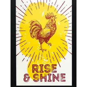 May include: A framed print with a yellow sunburst and a red rooster in the center. The text "RISE & SHINE" is below the rooster.