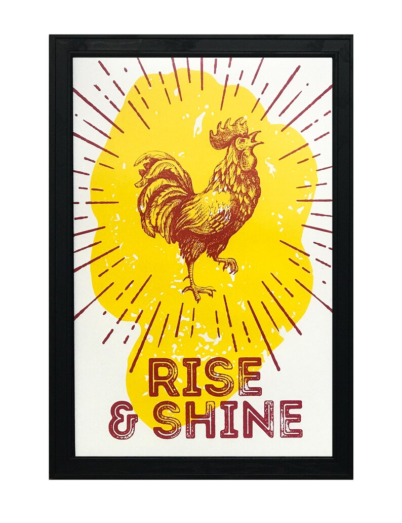 May include: A framed print with a yellow sunburst and a red rooster in the center. The text "RISE & SHINE" is below the rooster.