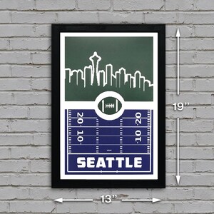 May include: A black framed print with a green and blue design featuring a skyline of Seattle, Washington. The print includes a football field with the word "SEATTLE" in white text. The print measures 13 inches wide by 19 inches tall.
