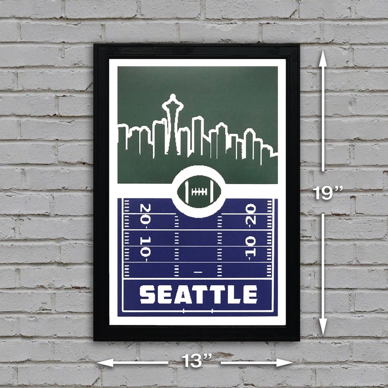 May include: A black framed print with a green and blue design featuring a skyline of Seattle, Washington. The print includes a football field with the word "SEATTLE" in white text. The print measures 13 inches wide by 19 inches tall.
