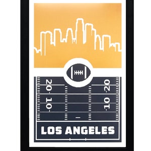 May include: A framed print with a gold background featuring a white silhouette of the Los Angeles skyline. The bottom of the print shows a black and white football field with a football in the center and the text "LOS ANGELES" below.
