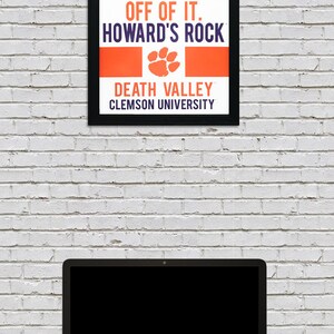 May include: A black framed poster with orange and purple text that reads "Give me 110% or keep your filthy hands off of it. Howard's Rock. Death Valley. Clemson University". The poster has a paw print graphic in the center.