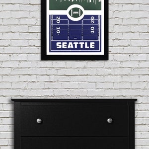 May include: A framed print of a football pitch with a skyline of Seattle, Washington. The skyline includes the Space Needle. The pitch is blue and the skyline is green. The text "SEATTLE" is printed below the pitch.
