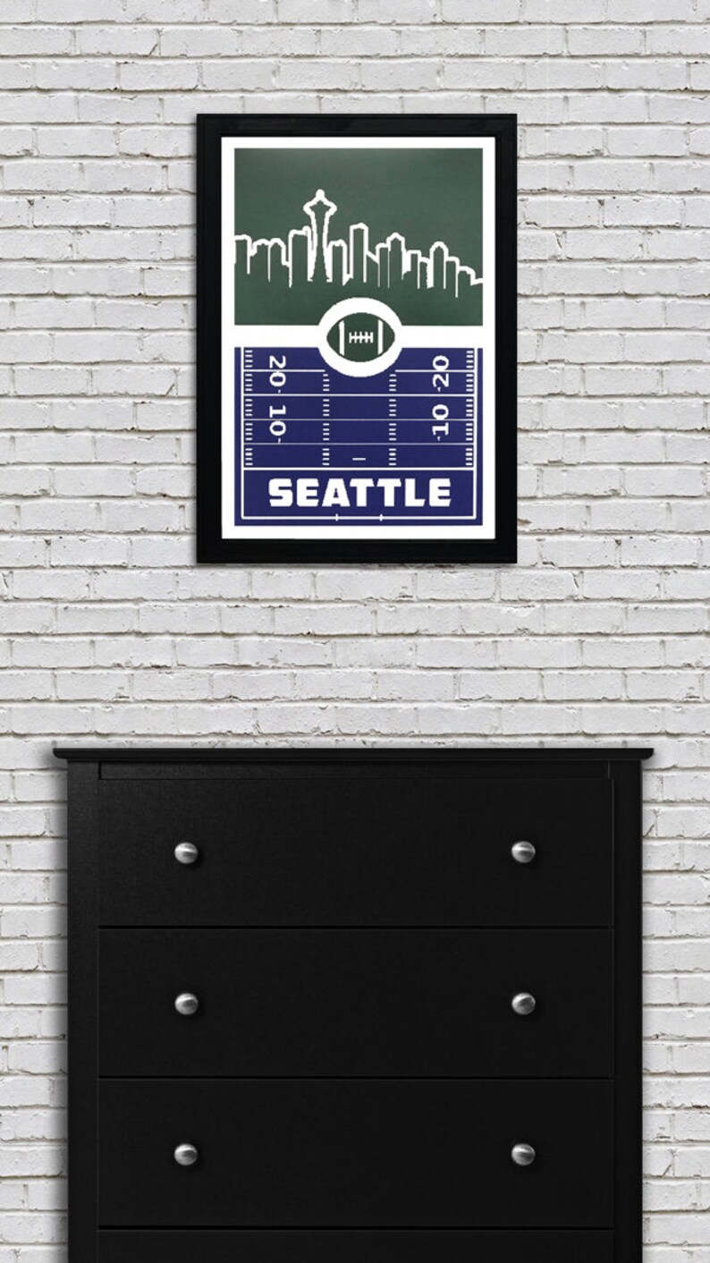 May include: A framed print of a football pitch with a skyline of Seattle, Washington. The skyline includes the Space Needle. The pitch is blue and the skyline is green. The text "SEATTLE" is printed below the pitch.