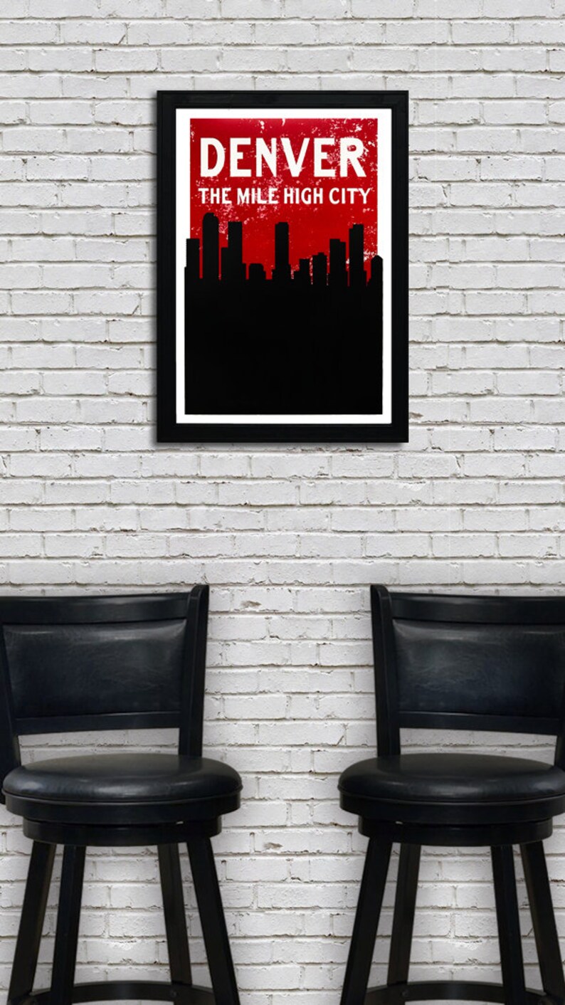 May include: A framed art print featuring the Denver skyline in black against a red background, with the city name in white. The text reads "DENVER THE MILE HIGH CITY." Below, two black bar stools are positioned in front of a white brick wall.
