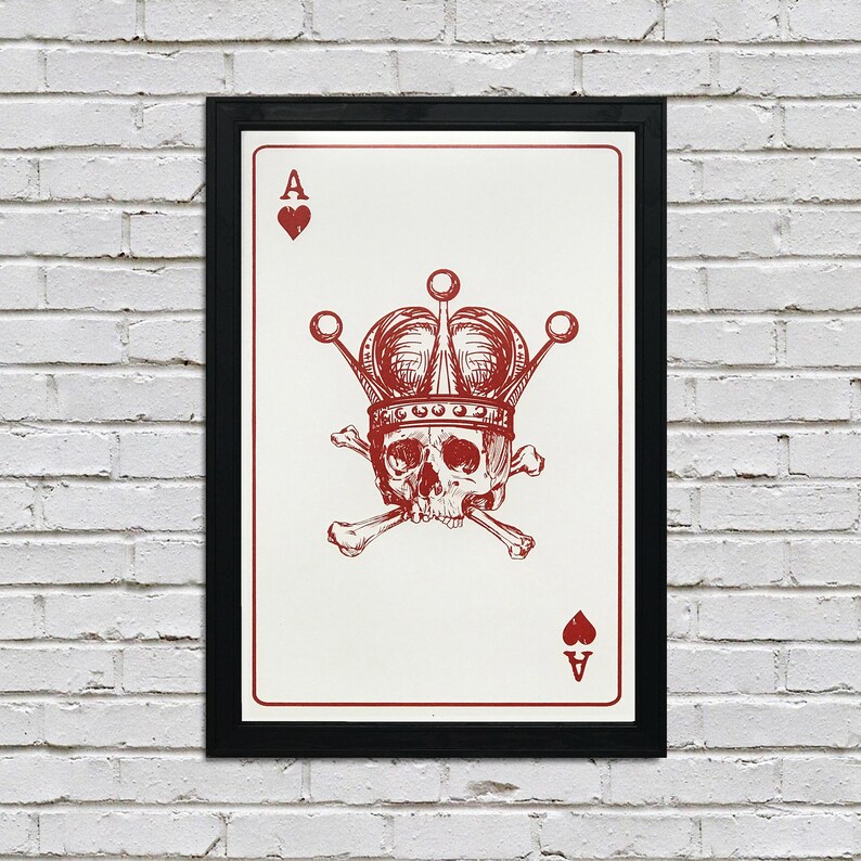May include: Framed art print featuring a red and white Ace of Hearts playing card design. The card depicts a skull wearing a crown, with crossed bones beneath it. The artwork is set against a white background and framed in black.