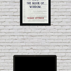 May include: A framed print with the quote "Honesty is the first chapter [in] the book of wisdom" by Thomas Jefferson. A laptop computer sits on a desk below the framed print, against a white brick wall.