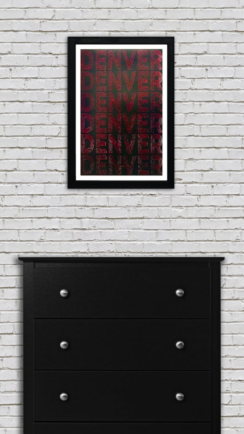 May include: A framed print with the word "DENVER" repeated five times in a red, white, and blue color scheme. The background is black with a subtle galaxy-like pattern.