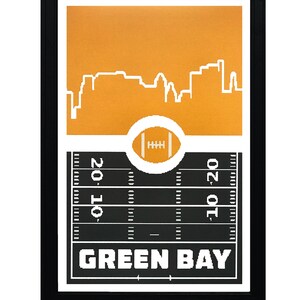 May include: A framed print with a black frame and a gold background. The print features a white silhouette of the Green Bay skyline with a football field and a football in the center. The text "GREEN BAY" is printed in white at the bottom of the print.