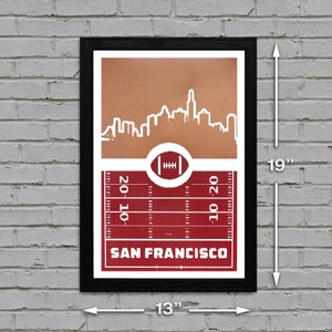 May include: A framed print with a red and brown color scheme. The print features a white silhouette of the San Francisco skyline with a football field and the text "SAN FRANCISCO" below. The print measures 13 inches wide by 19 inches tall.