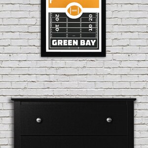 May include: A black and white framed print of a football field with a yellow silhouette of the Green Bay skyline. The text "GREEN BAY" is printed below the field.