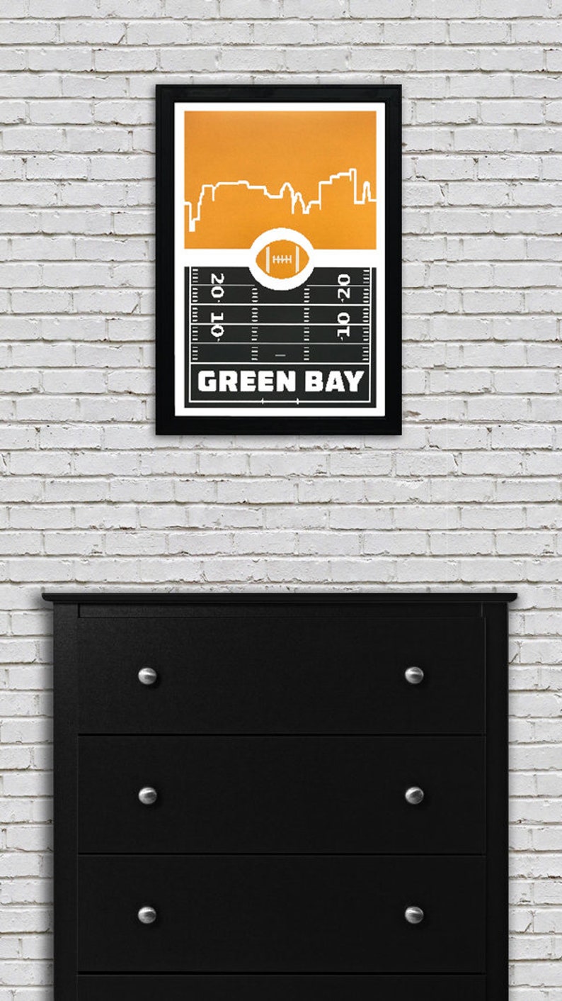 May include: A black and white framed print of a football field with a yellow silhouette of the Green Bay skyline. The text "GREEN BAY" is printed below the field.