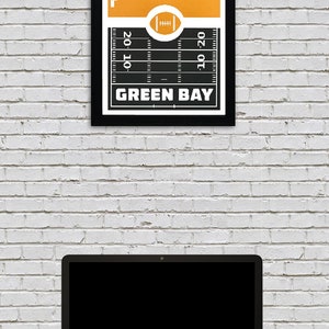 May include: A black framed print with a yellow background and a white silhouette of the Green Bay skyline. The print features a football field with the words "Green Bay" at the bottom.