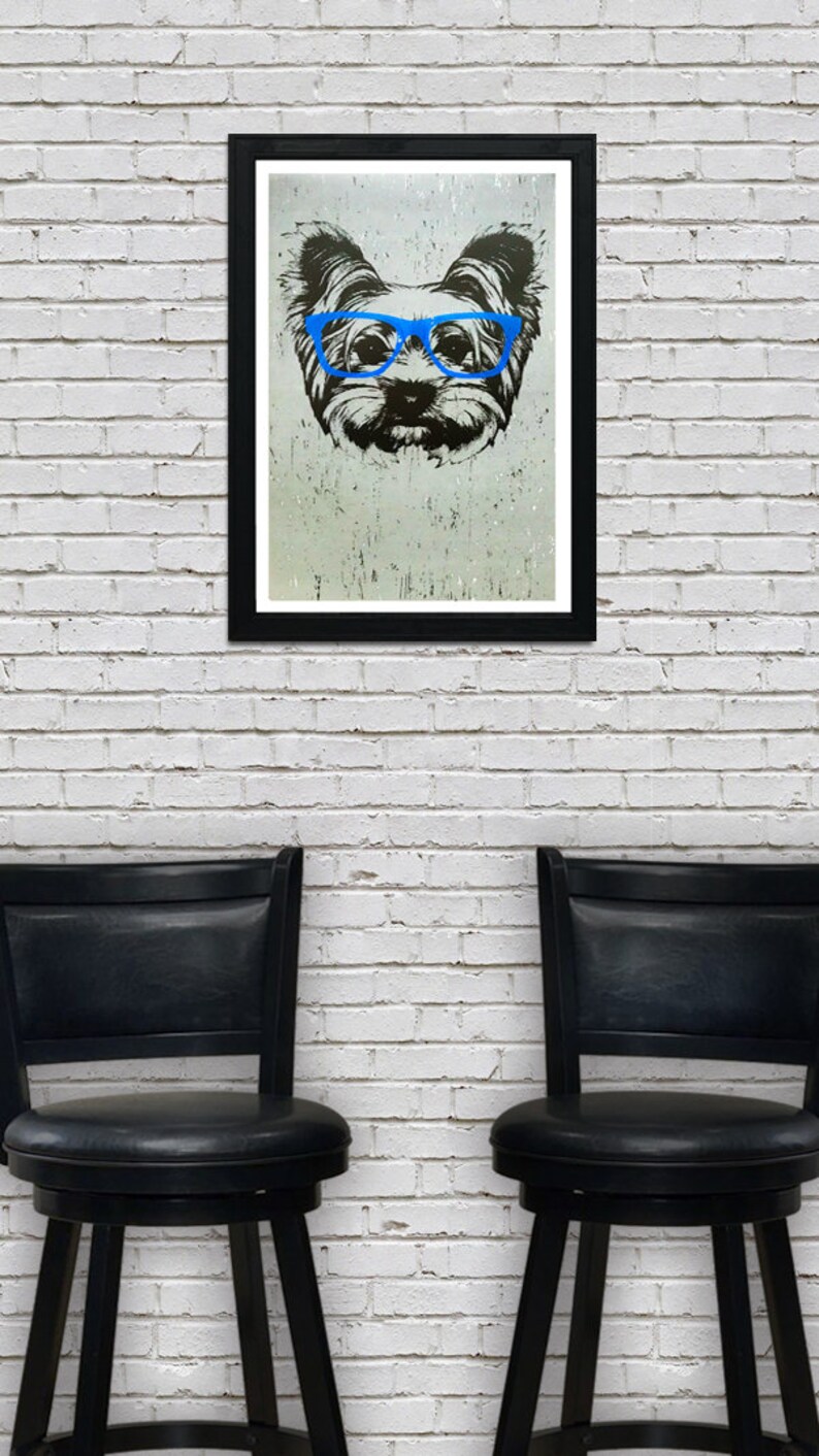 May include: A black framed print of a dog wearing blue glasses. The dog is a Yorkshire Terrier and the print is on a white brick wall background. Two black bar stools are in the foreground.